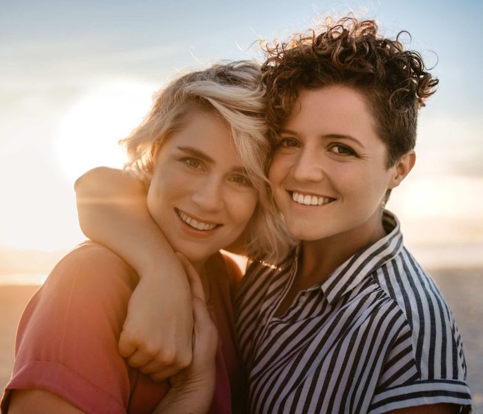 Portrait of a smiling young lesbian couple standing arm in arm together on a sandy beach at sunset